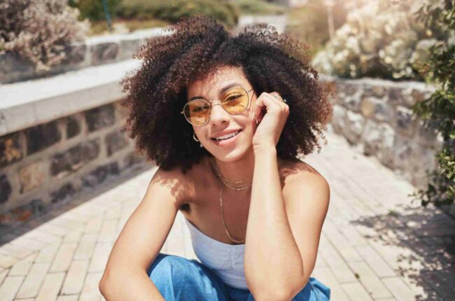 Mulher jovem com cabelos cacheados e óculos de sol sorrindo em um ambiente ao ar livre. A iluminação natural destaca sua beleza e estilo. Ideal para temas de moda e verão.